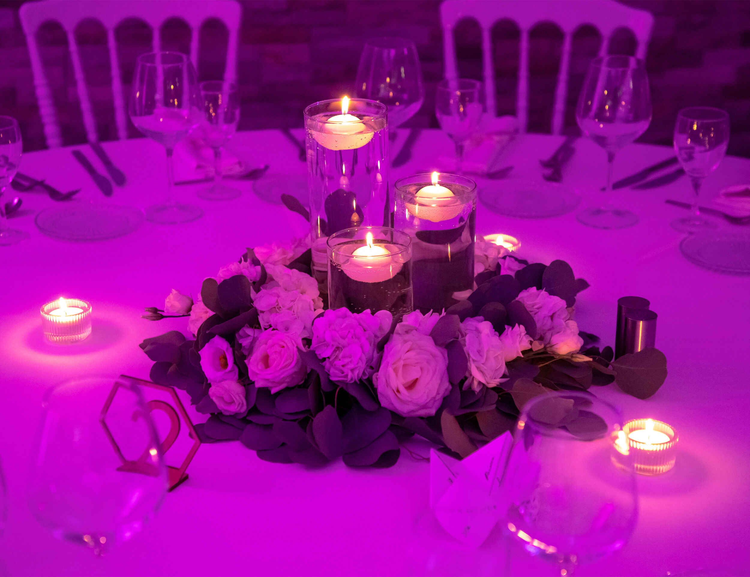 Romantic wedding table setting with pink lighting, floating candles, and a floral centerpiece surrounded by elegant glassware.